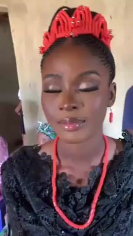 A young woman with dark skin is shown from the chest up. She is wearing a black lace top, a red coral necklace and earrings, and a red beaded crown. Her hair is styled in neat braids. She is wearing makeup, including eye shadow, eyeliner, and lip gloss. She closes her eyes, then opens them again. An older woman with a headwrap is partially visible in the background. The wall behind the young woman is plain and beige.