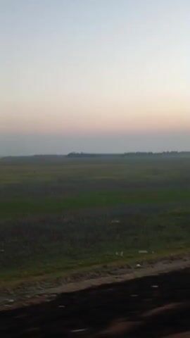 The video shows a landscape with a vast, flat field of green and brown under a pale sky. The perspective suggests movement, possibly from a vehicle, as evidenced by motion blur in the foreground and the appearance of a red object and a rearview mirror. The horizon is marked by a thin line of trees or structures, and the overall lighting suggests either dawn or dusk. There are no people or animals clearly visible in the frame. The environment appears rural and open.