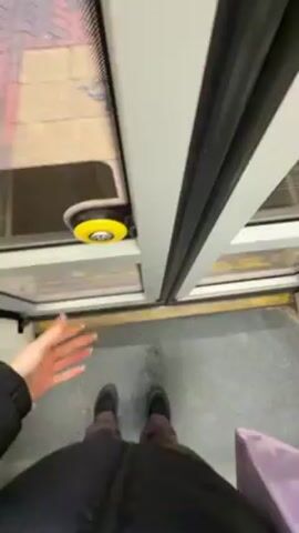 A person stands in the entryway of a light rail or subway car, facing a set of sliding doors that are slightly ajar. The person is wearing dark pants, a black jacket, and dark shoes. They are also holding a light purple shopping bag. A yellow and black button is visible on the door frame, presumably to open the doors fully. The person touches the button with their hand, possibly to open the doors further. Beyond the open doors, a red brick pattern is visible on what appears to be an exterior sidewalk. The floor inside the car is gray with a yellow safety line. The lighting is artificial and even.