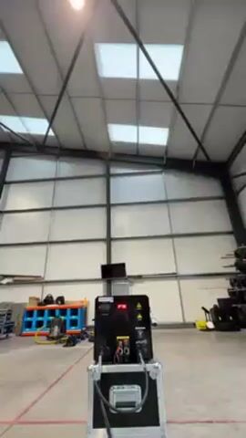 The video shows a pyrotechnic machine inside a large warehouse. The machine, enclosed in a black case, is in the center of the frame. It emits flames in a controlled manner. Initially, a straight stream of fire shoots upwards. Then, the machine emits several fireballs into the air. The warehouse has a high ceiling with square skylights and metal support beams. Various equipment and storage units are visible in the background. There are no people present.