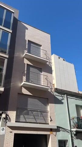 A low-angle video pans across a street scene featuring residential buildings under a clear blue sky. On the left, a tall, modern building with a light brick facade and three balconies with closed blinds is visible. Adjacent to it is a smaller, two-story building painted in a pale green, with a balcony featuring potted plants and a black railing. To the right of the green building, a row of older, attached houses with beige and yellow facades are seen. These houses have balconies with ornate railings and closed shutters on the windows. One of the balconies on a beige building has flowers and a metal railing. Further down the street, taller buildings are visible in the distance, along with trees and power lines. A dark-colored SUV is parked in the foreground, partially obscuring the view of the street and buildings on the right. A red Smart car is parked behind the SUV. The scene appears to be a sunny day in an urban or semi-urban environment.