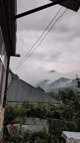 The video shows a view of misty mountains shrouded in clouds under an overcast sky. In the foreground, there is a house with a metal roof and siding. There are trees and vegetation in front of the house, as well as a table covered with a patterned tablecloth. Power lines are visible stretched across the frame. The scene suggests a rural or mountainous location, possibly experiencing inclement weather.