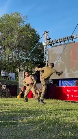 Two men in military fatigues are demonstrating martial arts on a grassy field. They are in front of a large screen showing a soldier saluting. Other people in military fatigues are sitting and kneeling nearby. Some civilians are standing behind a low stone wall watching the demonstration. There is a tree to the left, stage equipment and a red barrier to the right. The sky is blue. The two men grapple, and one man kicks the other, sending him to the ground.