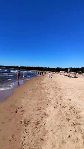 The video pans across a sandy beach on a clear, sunny day with a deep blue sky. Several people are scattered across the beach, some walking near the water's edge, others sunbathing or sitting on towels. In the background, a line of trees forms the edge of the beach. A building with large windows is visible in the distance. Several bicycles are parked on the sand. Some people are wearing swimwear. The overall scene depicts a relaxed atmosphere on a crowded beach.