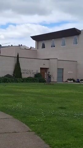 The video shows a low-angle shot of Parroquia de Santiago de Arteixo, a modern church in Arteixo, Spain. The church is a beige concrete structure with a low, flat roof and a simple bell tower. There is a statue of a woman and child in front of the church, and a grassy lawn with daisies. The sky is cloudy. A pedestrian walks on a path near the right corner of the building.