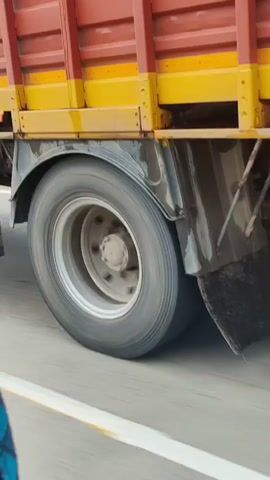 A close-up, low-angle shot of the rear wheel and mud flap of a large truck. The truck's tires are black and appear to be in motion, as suggested by the slight blur and the white line on the road surface. The mud flap is also black and textured, with some dirt or debris visible on its lower edge. The truck bed, made of red metal slats and yellow supports, is visible above the wheel. The background consists of a gray, out-of-focus road surface. The lighting is bright and natural.