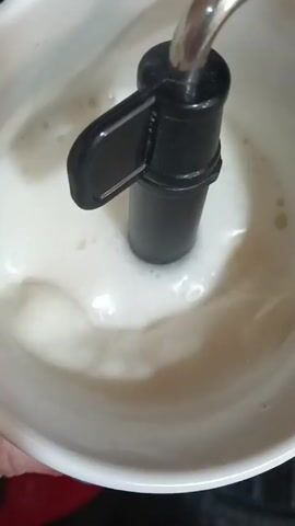 The video shows a top-down close-up of a white bowl containing milk being frothed by a milk frother. The frother, consisting of a chrome tube and a black plastic handle, is immersed in the milk. Bubbles and foam are visible in the milk as the frothing process continues. The bowl appears to be held by a hand, which is partially visible. The background is out of focus, with hints of red and black surfaces. Audio of background noise or indistinct voices is audible.