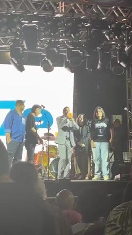 A group of five people stands on a stage in front of a large white screen displaying a blue logo that appears to be the number "39". A man in a light grey suit and purple tie holds a microphone and speaks, gesturing with his right hand. To his left, a woman in a black jacket and a woman in a dark t-shirt with a graphic design stand with their arms crossed. To the far left, a man in a blue shirt and a man in a black t-shirt are partially visible. Behind the speakers, a drum set is visible, and stage lighting is mounted overhead. The audience is blurred in the foreground.
