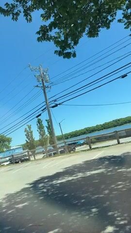 The video shows a drive-by of a waterfront establishment on a clear, sunny day.  Power lines and a utility pole are prominent in the foreground.  A parking lot is visible with some cars parked.  Beyond the parking lot, a wooden fence runs along the water's edge.  Several tall, slender trees stand behind the fence.  In the distance, a body of water and a tree-lined shore are visible.  The camera then pans to show the building itself, which has several kayaks mounted on its roof, each with a goose-like ornament. Signs on the building indicate services like "RENTALS" and "LESSONS."  An American flag is visible.  There are people visible near a dark-colored SUV in the parking lot.  Green foliage and flowers are present in the foreground.  A dark pickup truck is also briefly visible.