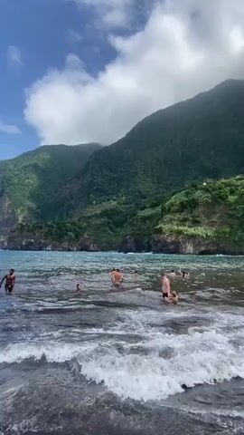 The video shows a black sand beach with people swimming and walking in the shallow ocean water. In the background, a steep, green, terraced mountain rises dramatically from the sea. Buildings are visible on the mountain slopes. The sky is partly cloudy. The waves are moderate, with white foam washing onto the shore. People of various ages are scattered throughout the water and on the beach.