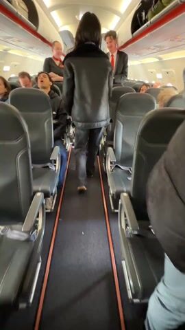 The video shows the aisle of a passenger airplane cabin, between rows of black seats with orange accents. A person wearing a black leather jacket and dark pants walks towards the front of the plane, passing a flight attendant in a black uniform with orange trim and a male flight attendant in a suit with a red tie. Passengers are seated on either side of the aisle, some looking towards the front. The lighting is bright, and the interior of the plane appears clean and modern. The camera follows the person walking down the aisle.