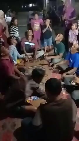 A group of people, including children and adults, are sitting on the ground in a semi-circle at night, participating in a game. Some of the adults have small objects, likely crackers or biscuits, balanced on their foreheads. They appear to be attempting to move the objects into their mouths without using their hands. Several children are standing and observing the game. The environment suggests an outdoor gathering, possibly a street or yard, with some artificial lighting visible in the background. The overall atmosphere seems festive.
