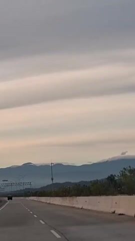 The video shows a highway with a grey sky and rolling clouds. Mountains are visible in the distance, partially obscured by mist. Trees and palm trees line the side of the road. A few cars are on the highway, and there are some overhead structures, possibly for tolls or signage. The camera is positioned in a moving vehicle, likely at eye level. The overall atmosphere is serene and possibly captures dawn or dusk.