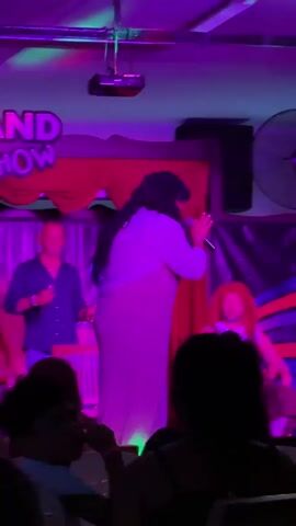 A stage performance featuring two individuals. One individual, dressed in a light-colored dress and dark wig, is actively singing into a microphone. The stage is adorned with a banner that reads "GRAND SHOW" in large lettering. The lighting changes dynamically, cycling through blue, green, red, and purple hues. In the background, another person, appearing to be a male performer with a bald head, is also on stage, gesturing with their arms. The audience is visible in the foreground, seated and watching the performance.