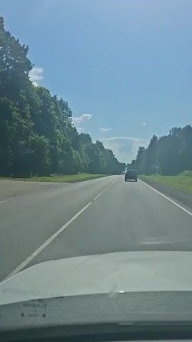 The video shows a POV shot from inside a vehicle driving on a paved road. The road has two lanes, and the vehicle is in the right lane. A single car is visible ahead in the left lane. Both sides of the road are lined with trees. The sky is blue with a few white clouds, and the sun appears to be shining brightly. The video is taken during the daytime.
