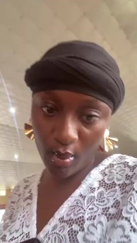 The video shows a close-up of a woman wearing a black headscarf, large gold earrings, and a white lace top with a colorful patterned fabric underneath. She makes faces and gestures towards the camera. In the background, another woman wearing an orange tie-dye top is visible. The environment appears to be indoors with a white ceiling with lights.