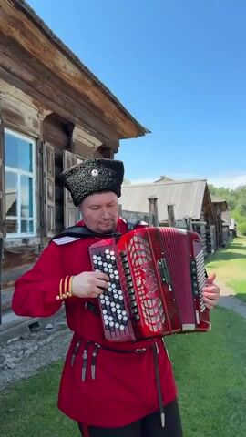 A man in traditional Cossack attire, including a red tunic, black hat, and gold armbands, plays a red accordion. He stands outdoors in front of a rustic wooden building with shuttered windows. The accordion has white and black keys on the left side and a bellows with decorative red patterns on the right. The man is focused on playing the instrument. The sky above is clear and blue.