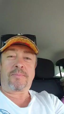 The video captures a selfie-style shot from inside a vehicle. The camera is held by a man wearing a white t-shirt and a patterned baseball cap. He is in the foreground and looking towards the camera with a slight smile. Behind him, in the passenger seat, a woman with blonde hair is visible. She is wearing a striped top in pink, white, and blue colors and is also looking towards the camera and smiling. The interior of the car is visible, including headrests and the ceiling. The overall impression is a casual, personal moment being recorded.
