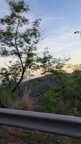 The camera is moving along a road. The road has a guardrail on the side. Beyond the guardrail is a forest. Behind the forest are mountains. Above the mountains is a blue sky with some clouds and orange light from the sunset. A few trees obstruct the view.