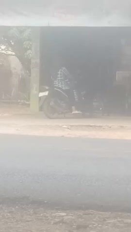The video shows a person wearing a checkered shirt and pants sitting on a motorcycle in front of an open-air structure. The person is in the process of starting or operating the motorcycle. Dust is being kicked up around the motorcycle and the ground. The environment appears to be outdoors with a road in the foreground. The lighting is somewhat hazy or overexposed.