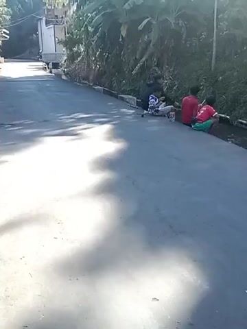 A group of young males are sitting on the side of a paved road. Some are sitting directly on the asphalt, while others are sitting on the curb next to a small drainage ditch. They appear to be engaged in some activity or conversation. There are some bags and containers near them. The road is lined with vegetation on one side, and there are some buildings and a vehicle in the background. The sun is shining, casting shadows on the road. One individual on a motorcycle passes by in the latter part of the video.