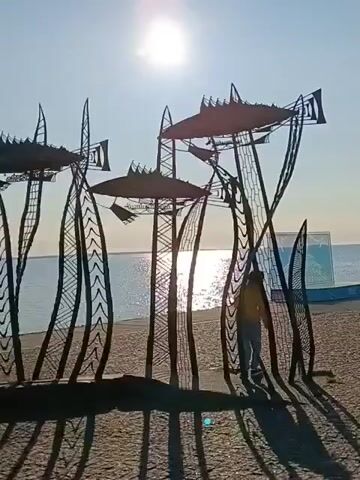 The video shows a beach scene with a metal sculpture in the foreground. The sculpture consists of several tall, abstract plant-like structures with fish silhouettes at the top, appearing as if they are swimming through seaweed. The sun is shining brightly, casting long shadows on the sandy beach. The ocean is visible in the background. A person, wearing a yellow hoodie and blue jeans, is standing to the right of the sculpture, partially obscured by one of the metal elements. The person appears to be interacting with the sculpture, possibly trying to go through it, but then stops. Another person is partially visible on the left side of the sculpture. The overall atmosphere is bright and sunny, with the sounds of the ocean and speech.