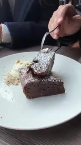 The video shows a close-up of a dessert being split with a spoon. The dessert is a dark brown chocolate brownie, square-shaped, dusted with powdered sugar. Next to it is a scoop of white ice cream, sprinkled with crushed nuts. A person wearing a black jacket and a white-sleeved shirt is holding the spoon. The dessert is on a white plate, set on a brown, textured surface. The focus remains on the brownie being divided.