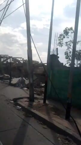 This video shows a street view of a destroyed building. The building is in ruins, with rubble and debris scattered everywhere. The roof appears to be partially collapsed, and some walls are still standing but are heavily damaged. In the foreground, there are utility poles and wires, suggesting the location is an urban or suburban area. The sky is cloudy, and the lighting suggests it is daytime. There are no people visible in the video.