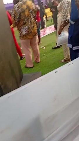 The video shows a crowd of people at an outdoor event on a grassy area. Many are holding up phones, likely taking pictures or videos. Some people are wearing red shirts. There's a DJ booth in the foreground with a large speaker. In the background, there's a decorative structure and a white tent. There are wads of cash on the ground near the people wearing red.