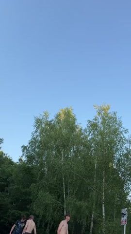 The video displays a clear blue sky over a lush green forest. In the foreground, several people are enjoying a day at a lake. Some are swimming in the water, while others are relaxing on the sandy shore. A few individuals are seen on paddleboards, indicating a leisurely outdoor activity. The overall atmosphere appears to be one of recreation and relaxation.