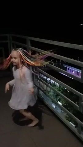 The video shows a young girl with colorful braided hair dancing on a balcony at night. She is wearing a light-colored dress and is barefoot. The balcony has a metal railing, and behind it, one can see lights from buildings and trees, suggesting an urban or resort setting. The girl is in motion, her arms and body creating a blurred effect due to the movement. The overall mood is energetic and playful. The time of day is clearly night, indicated by the dark sky and artificial lighting. There is no visible text in the video. The location is identified as Gagra, Abkhazia.