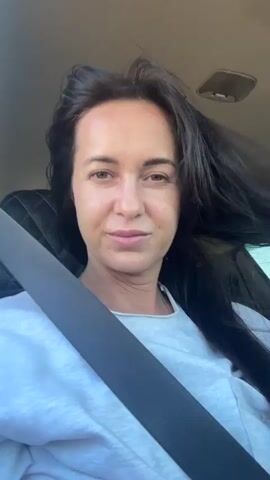 A woman with long, dark hair is in the passenger seat of a car, wearing a light gray sweatshirt and a black seatbelt. Her hair is being blown by the wind, and she is smiling at the camera. The background shows the interior of the car, including a black headrest and a window.