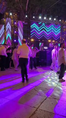 The video shows an outdoor party at night.  There is a dance floor lit with colorful lights, predominantly blue, purple, and green.  A large LED screen in the background displays a zig-zag pattern of purple and green lights.  Several people are dancing, most of them men. Some are wearing yarmulkes.  There are trees and decorative lights visible in the background.  A DJ booth with equipment is set up to the right of the LED screen.  The overall atmosphere appears festive.