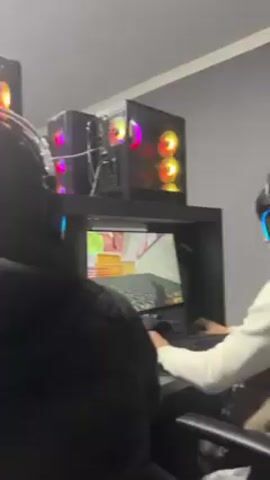 The video shows a dimly lit room with two individuals seated at computer stations playing a video game. Each station has a computer monitor displaying the game, a keyboard, and a mouse. The computer towers are visible, each with colorful LED lights. One individual is wearing a headset with blue lights. The other person is wearing a white sweatshirt. The focus shifts between the two individuals as they play the game.