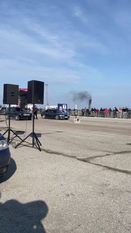 The video shows two vehicles participating in a drag race event. A gray Volkswagen Golf Mk2 and a black Mercedes-Benz W124 are positioned side by side at the starting line, and begin to accelerate, creating a large cloud of black smoke.  A crowd of people is standing behind a barrier along the edge of the track, watching the race. There are loudspeakers and flags alongside the track. The sky is blue with scattered clouds, and the race track is an asphalt surface with faded lane markings. The vehicles speed away from the starting line, kicking up dust as they go.