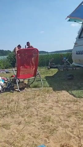 A sunny day at a beach with a lake in the background. Several people are visible on the sand, some lying on towels and others sitting. In the foreground, a red folding chair is positioned near a group of people. Two individuals, one in a black bikini and another in red swim trunks, are standing near the chair. In the lake, several people are swimming or on inflatable devices. The shoreline is lined with trees.