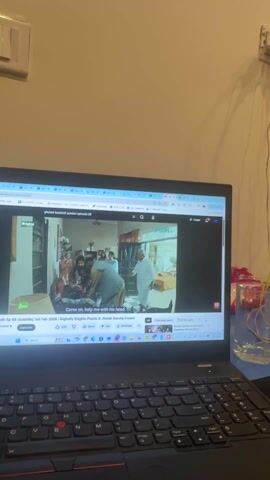 The video shows a laptop displaying a YouTube video titled "ghulum badshah sundari episode 09." The screen shows a scene with multiple people in an interior setting. The laptop sits on a desk with a glass of water and a packet of sweets next to it. A wall is visible in the background, with an electrical outlet and wires.