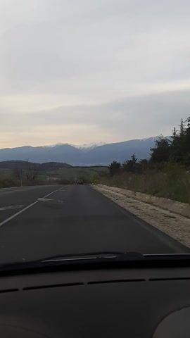 From inside a vehicle traveling on a road, we see a view of a mountain range in the distance with snow-capped peaks. A car is visible on the road ahead, driving in the same direction. The sky is overcast with pale clouds. The roadside is lined with green vegetation and trees. The video appears to be taken at dusk.