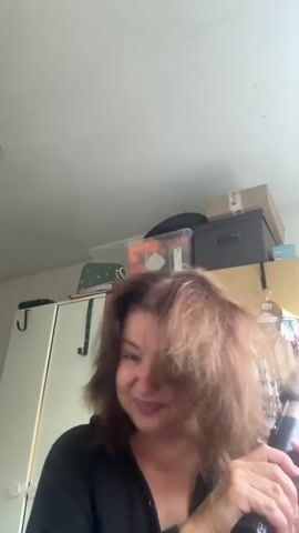 In this video, a woman with medium-length, reddish-brown hair is shown in an indoor setting, likely her home in Steenwijk, Netherlands. She is holding a black and gold hairdryer and appears to be in the process of drying her hair. Her hair is being blown around by the hairdryer, making it look somewhat messy and voluminous. She is wearing a black top.

The background reveals a cluttered shelf with various boxes and containers, suggesting a storage area. A white cabinet is visible on the left side of the frame, and a piece of clothing, possibly a patterned shirt, hangs on a rack to the right. The lighting suggests it is daytime, with natural light coming from above.

The woman's facial expressions shift throughout the short clip. Initially, she seems to be struggling slightly with her hair, with a slight smile. In later frames, her smile widens, and she appears to be enjoying herself, perhaps finding humor in the activity. The overall mood is lighthearted and informal. No visible text is present in the media file.