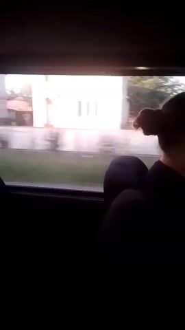The video shows a view from inside a vehicle, looking out the passenger window. The window frames a rural landscape with trees and fields. A person's head and shoulder are visible in the foreground, looking out the window. The person has dark hair pulled back into a bun. The vehicle appears to be in motion, as indicated by the blurred scenery outside. The time stamps indicate the video is a short clip.