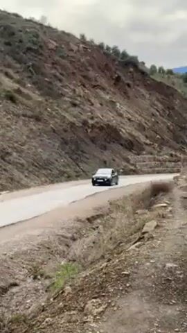 A black car drives on a paved road through a mountainous, dry landscape. The road winds through the terrain, with hillsides covered in sparse vegetation on either side. The sky is overcast, with the sun peaking through the clouds. The camera angle changes slightly, panning upwards to follow the car as it moves along the road. There are rocks and dirt in the foreground, adding to the ruggedness of the scene.