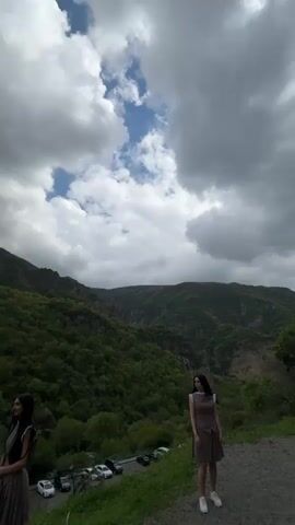 The video shows a group of young women in brown dresses standing in front of a mountainous landscape on a cloudy day. Several cars are parked nearby. As the camera pans, more people are seen walking around, some taking pictures. There is a coin-operated telescope set up as well. The landscape is primarily green hills with rocky outcroppings under a sky of gray clouds punctuated by patches of blue.