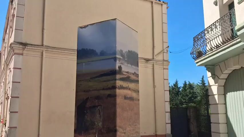 The video shows a rectangular information pillar, titled "Canadal", standing on a paved surface next to a building. The pillar has an image of a landscape with a lake on its top section and textual information on the lower sections. The text appears to be in Spanish or Catalan, along with a map and a QR code. The buildings surrounding the pillar appear to be European-style architecture, with a balcony featuring ornate metalwork on one. The weather appears sunny, with the shadows of trees visible on the ground.