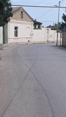 A black SUV is captured driving down a residential street. The road surface is asphalt and shows tire marks from previous maneuvers. Buildings with white walls and stone accents line the street. The sky is overcast and a utility pole is visible. The vehicle is in motion and appears to be turning.