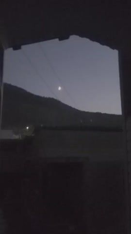 A video taken from inside a dark room shows a view of a crescent moon in a pale, dusk sky. Two power lines run diagonally across the frame, with the moon positioned between them. A dark, blurry landscape with a hill or mountain is visible in the background. Some faint lights, possibly from buildings, are visible on the left side of the landscape. The foreground is very dark, obscuring any details of the room. The video is short, with the camera slightly shaking throughout.