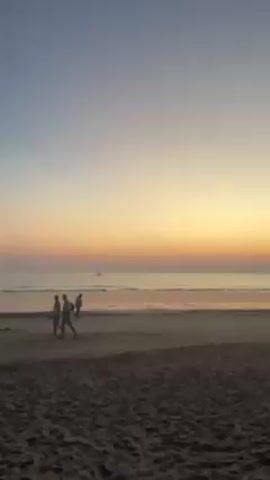 The video shows a beach at sunset. The sky transitions from blue to yellow and orange as it approaches the horizon. The ocean is calm, with small waves breaking on the shore. Several people are walking along the beach, appearing as silhouettes against the light. In the distance, there is a small boat on the water. The sand is dark in the foreground and reflects the light of the sunset.