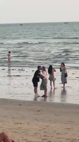 On a sandy beach with shallow water, a man and a woman embrace and kiss. Two women nearby take pictures with their phones. Another person wades in the water further back. The sky is overcast.  Several small boats are visible on the water in the distance.