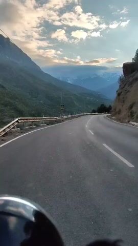 The video is a first-person perspective of a motorcycle ride on a paved road that curves to the right. The road is bordered by a metal guardrail on the left, and a steep, rocky incline with sparse vegetation on the right. Beyond the guardrail, lush green mountains rise sharply, covered in trees. In the distance, majestic, snow-capped mountains are visible under a cloudy sky with hints of blue. The road appears to be a mountain pass. The bottom of the frame shows a portion of the motorcycle's headlight or dashboard. The overall impression is of a scenic and potentially remote mountain landscape.