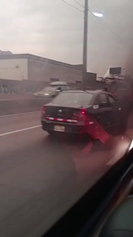 The video shows a vehicle moving on a road, as seen through its side window. Outside, a black sedan is visible on the left, followed by a red pickup truck passing on the right. Further ahead, a motorcycle moves along the road. The weather appears overcast or hazy. The reflection of the car's interior is visible in the window, including the viewer's arm and a portion of the seat.