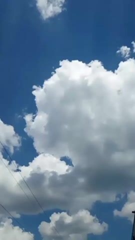 The video shows a bright blue sky with fluffy white clouds. There are power lines running across the sky. In the bottom right corner, a building can be seen. In the bottom left corner, there are trees and a utility pole. The camera appears to be moving, possibly in a vehicle, as evidenced by the shifting perspective of the clouds and the power lines.